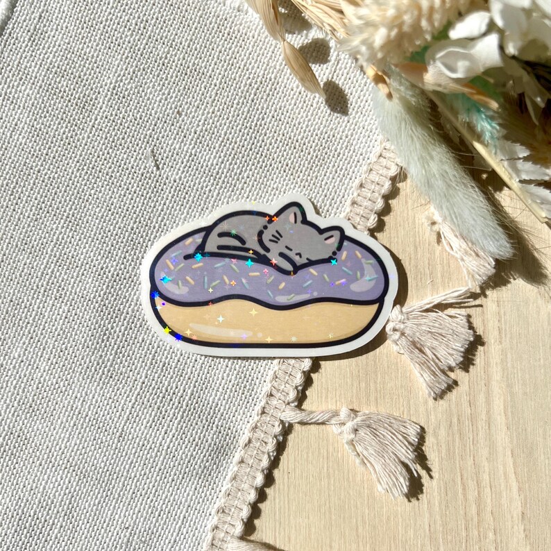 May include: A sticker featuring a gray cat sleeping on a purple frosted donut with sprinkles. The donut has a tan base and a white border. The sticker has a holographic effect.