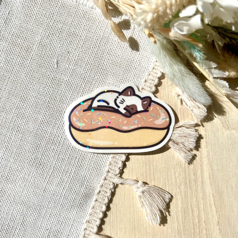 May include: A sticker featuring a cartoon cat sleeping on a donut. The donut has a light brown glaze with colorful sprinkles. The cat is white with brown markings and is curled up on the donut.