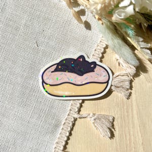 May include: A sticker featuring a black cat sleeping on a pink frosted donut with colorful sprinkles. The donut has a yellow base and a white outline. The sticker has a holographic effect.