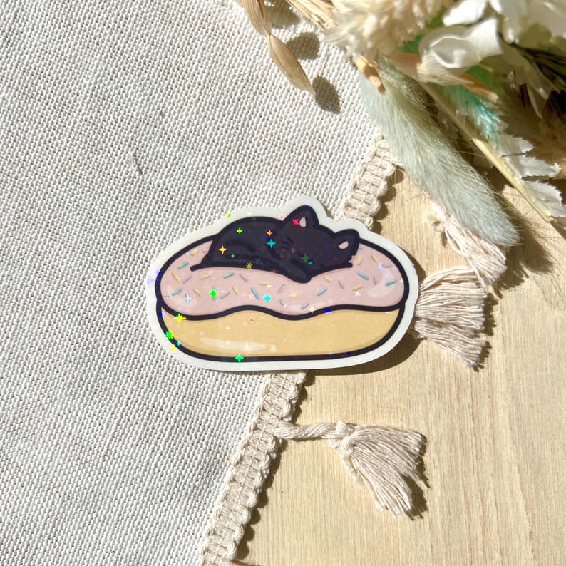 May include: A sticker featuring a black cat sleeping on a pink frosted donut with colorful sprinkles. The donut has a yellow base and a white outline. The sticker has a holographic effect.