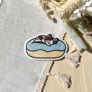 May include: A sticker featuring a sleeping cat on a blue donut with sprinkles. The cat has brown, black, and white fur. The donut has a gold outline with iridescent accents. The sticker is on a neutral background.