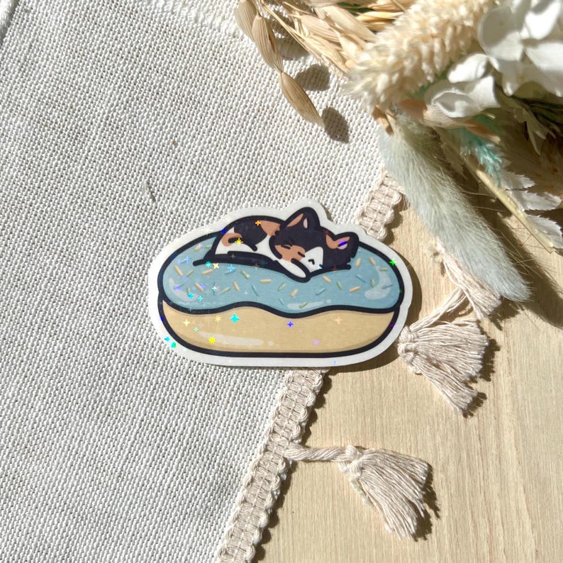 May include: A sticker featuring a sleeping cat on a blue donut with sprinkles. The cat has brown, black, and white fur. The donut has a gold outline with iridescent accents. The sticker is on a neutral background.