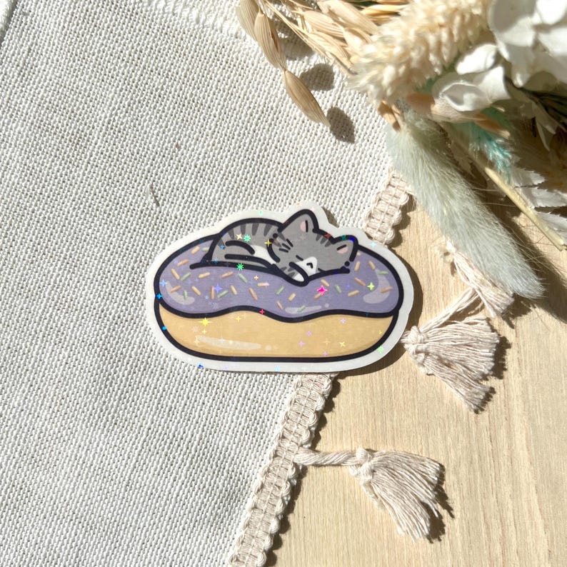May include: A sticker featuring a gray tabby cat sleeping on a purple frosted donut with colorful sprinkles. The donut has a yellow base and a black outline. The sticker is on a light-colored fabric surface.