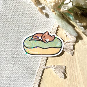 May include: A sticker featuring a cartoon cat sleeping on a green frosted donut. The donut has sprinkles and a brown outline. The cat is brown and white with its eyes closed. The sticker has a glittery, holographic effect.