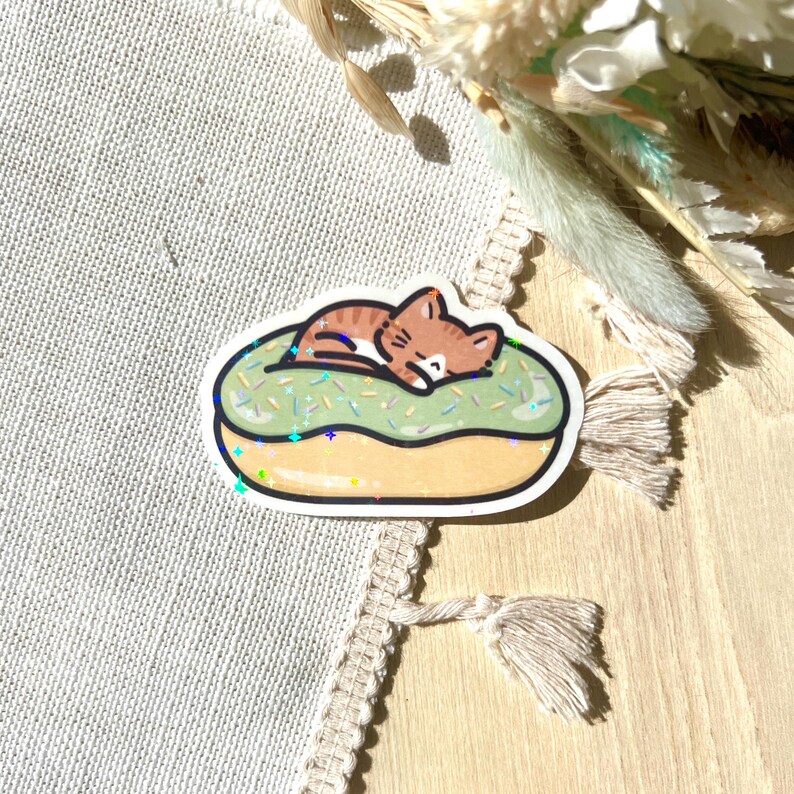 May include: A sticker featuring a cartoon cat sleeping on a green frosted donut. The donut has sprinkles and a brown outline. The cat is brown and white with its eyes closed. The sticker has a glittery, holographic effect.