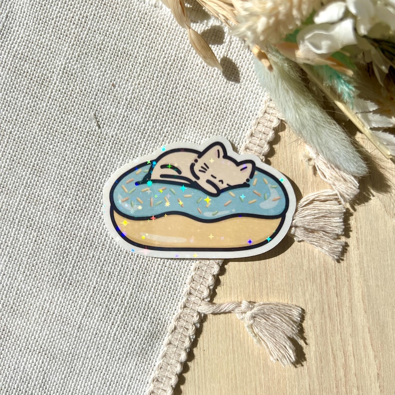 May include: A sticker featuring a cartoon cat sleeping on a blue donut with sprinkles. The donut has a light brown base and a light blue frosting with colorful sprinkles. The sticker has a holographic effect.