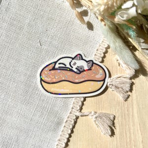 May include: A sticker featuring a cartoon cat sleeping on a donut. The donut has a light brown glaze with sprinkles. The cat is white with gray markings. The sticker has a white border and a glittery effect.