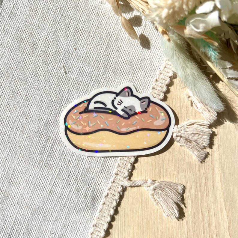 May include: A sticker featuring a cartoon cat sleeping on a donut. The donut has a light brown glaze with sprinkles. The cat is white with gray markings. The sticker has a white border and a glittery effect.