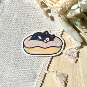 May include: A sticker featuring a black cat sleeping on a pink donut with sprinkles. The donut has a golden-yellow glaze and is outlined in white. The sticker has a glittery, holographic effect.
