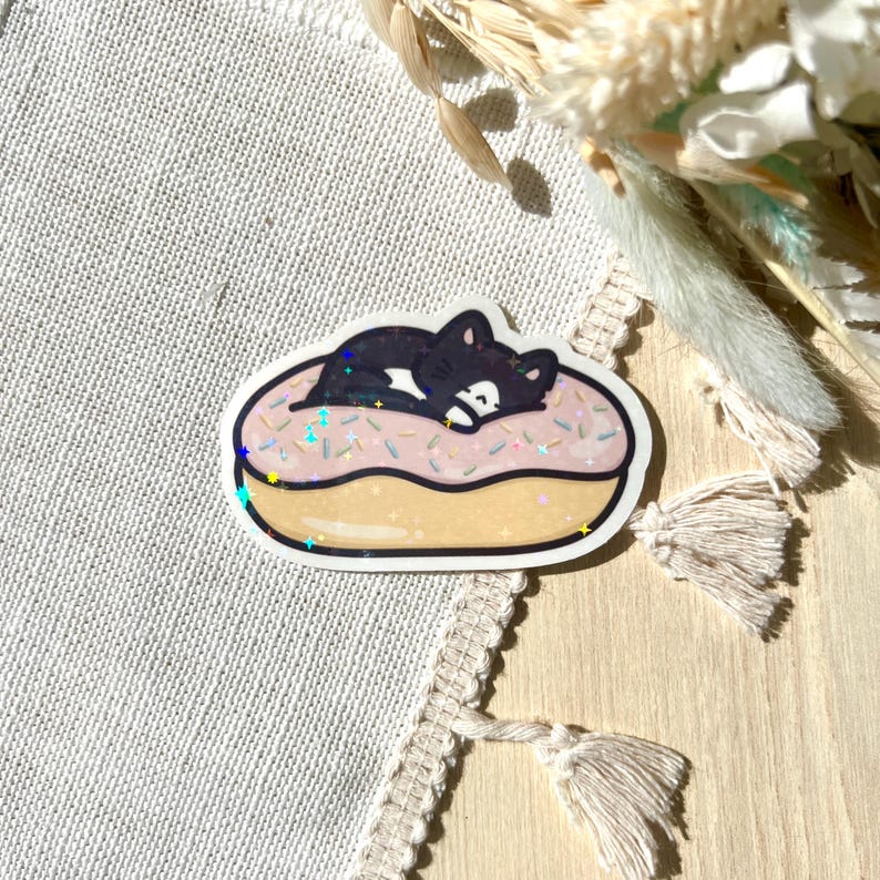 May include: A sticker featuring a black cat sleeping on a pink donut with sprinkles. The donut has a golden-yellow glaze and is outlined in white. The sticker has a glittery, holographic effect.
