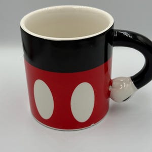May include: A ceramic mug with a design inspired by a famous cartoon character. The mug has a red base with two white ovals, a black upper section, and a black handle shaped like a hand. The interior is white.