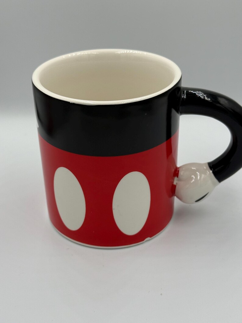 May include: A ceramic mug with a design inspired by a famous cartoon character. The mug has a red base with two white ovals, a black upper section, and a black handle shaped like a hand. The interior is white.