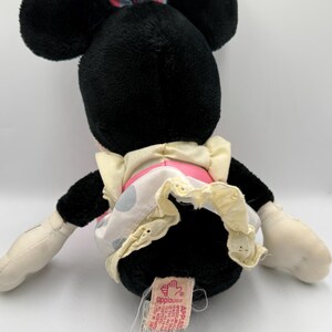 May include: A black plush Minnie Mouse doll with large black ears, wearing a light blue bow with pink polka dots and a pink and white polka dot dress with a cream-colored trim. The doll has white gloves and a tag that says "applause".