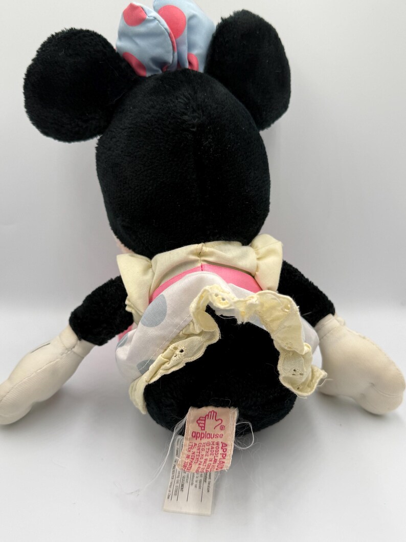 May include: A black plush Minnie Mouse doll with large black ears, wearing a light blue bow with pink polka dots and a pink and white polka dot dress with a cream-colored trim. The doll has white gloves and a tag that says "applause".