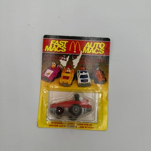 Puede incluir: Un coche de juguete rojo con una figura de conductor negro. El coche está en una tarjeta amarilla y blanca con las palabras "Fast Macs Auto Macs" impresas en ella. La tarjeta también tiene una imagen de otros cuatro coches de juguete.