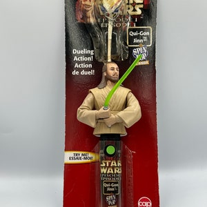 May include: A Star Wars Episode I Qui-Gon Jinn Spin Pop candy toy. The toy is a plastic figure of Qui-Gon Jinn holding a lightsaber. The figure is attached to a plastic stick with a green Spin Pop candy attached to the end. The packaging is red and features the Star Wars logo and the words "Episode I" and "Qui-Gon Jinn".