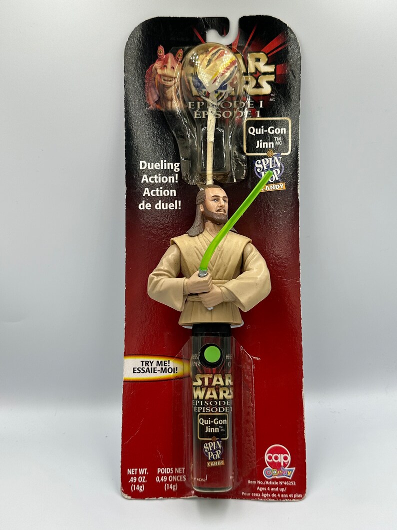 May include: A Star Wars Episode I Qui-Gon Jinn Spin Pop candy toy. The toy is a plastic figure of Qui-Gon Jinn holding a lightsaber. The figure is attached to a plastic stick with a green Spin Pop candy attached to the end. The packaging is red and features the Star Wars logo and the words "Episode I" and "Qui-Gon Jinn".