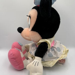 May include: A plush Minnie Mouse doll, seated and viewed from the side. The doll has a black head and ears, a pink and white polka dot bow, and a light yellow dress with blue polka dots. Her shoes are pink.