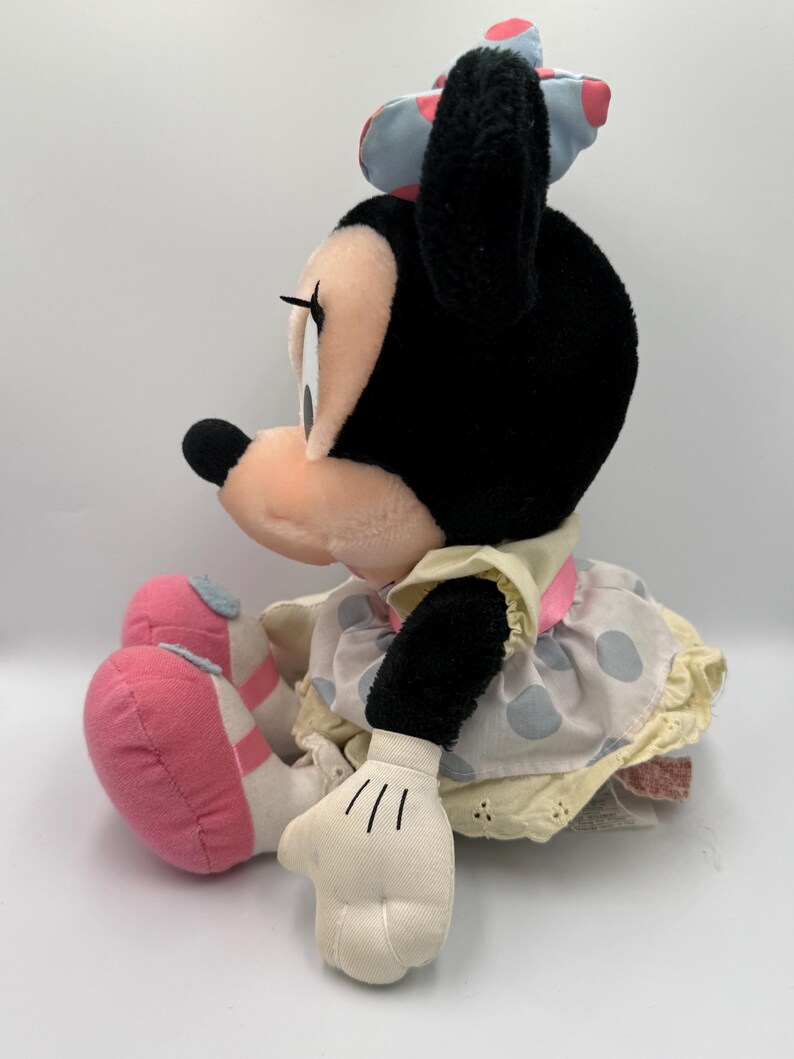 May include: A plush Minnie Mouse doll, seated and viewed from the side. The doll has a black head and ears, a pink and white polka dot bow, and a light yellow dress with blue polka dots. Her shoes are pink.