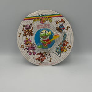 May include: A Jim Henson's Muppet Babies Stage Stars Baby Kermit pin on a circular cardboard backing. The pin features a green frog playing a yellow and red guitar. The backing displays illustrations of Muppet characters and musical notes.