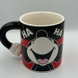 May include: A ceramic coffee mug with a cartoon character's smiling face and the word "HA" on either side. The mug has a black handle and a red and black design. The handle is shaped like a cartoon hand.