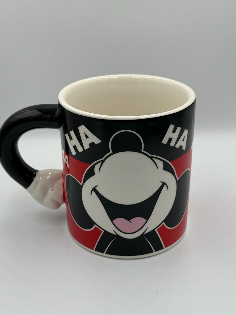 May include: A ceramic coffee mug with a cartoon character's smiling face and the word "HA" on either side. The mug has a black handle and a red and black design. The handle is shaped like a cartoon hand.
