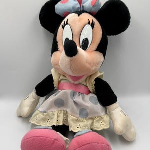 May include: A plush Minnie Mouse doll with black ears, a pink face, and large white eyes. She wears a light blue bow with pink polka dots, a cream-colored dress with gray polka dots, and pink shoes. The doll is soft and cuddly.