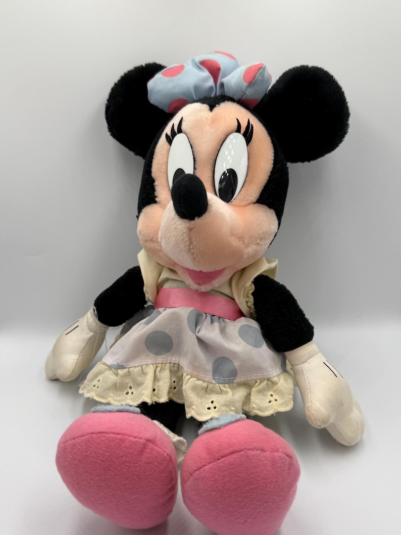 May include: A plush Minnie Mouse doll with black ears, a pink face, and large white eyes. She wears a light blue bow with pink polka dots, a cream-colored dress with gray polka dots, and pink shoes. The doll is soft and cuddly.