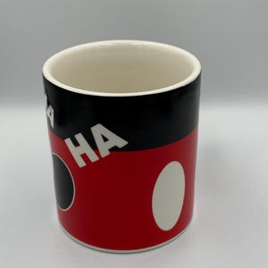 May include: A cylindrical ceramic mug with a white interior. The exterior features a black top section, a red mid-section with white oval and black circle accents, and the word "HA" in white.