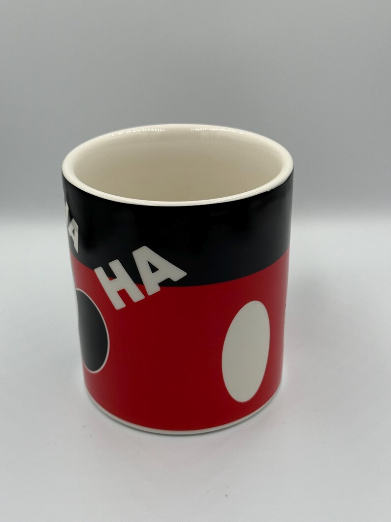 May include: A cylindrical ceramic mug with a white interior. The exterior features a black top section, a red mid-section with white oval and black circle accents, and the word "HA" in white.
