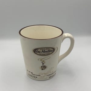 May include: A cream-coloured ceramic mug with a brown rim and handle. The front features the Tim Hortons logo with the text Always Fresh and Toujours Frais, a small steaming coffee cup icon, and the words coffee, cappuccino, and mocha printed.