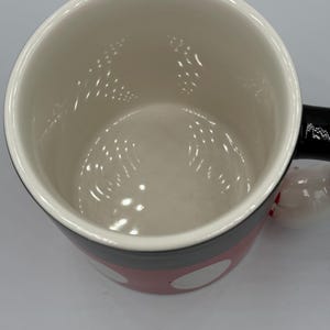 May include: A ceramic mug with a white interior and a red exterior decorated with white polka dots. The mug has a black handle and a black rim. The mug is designed with a cartoon theme.