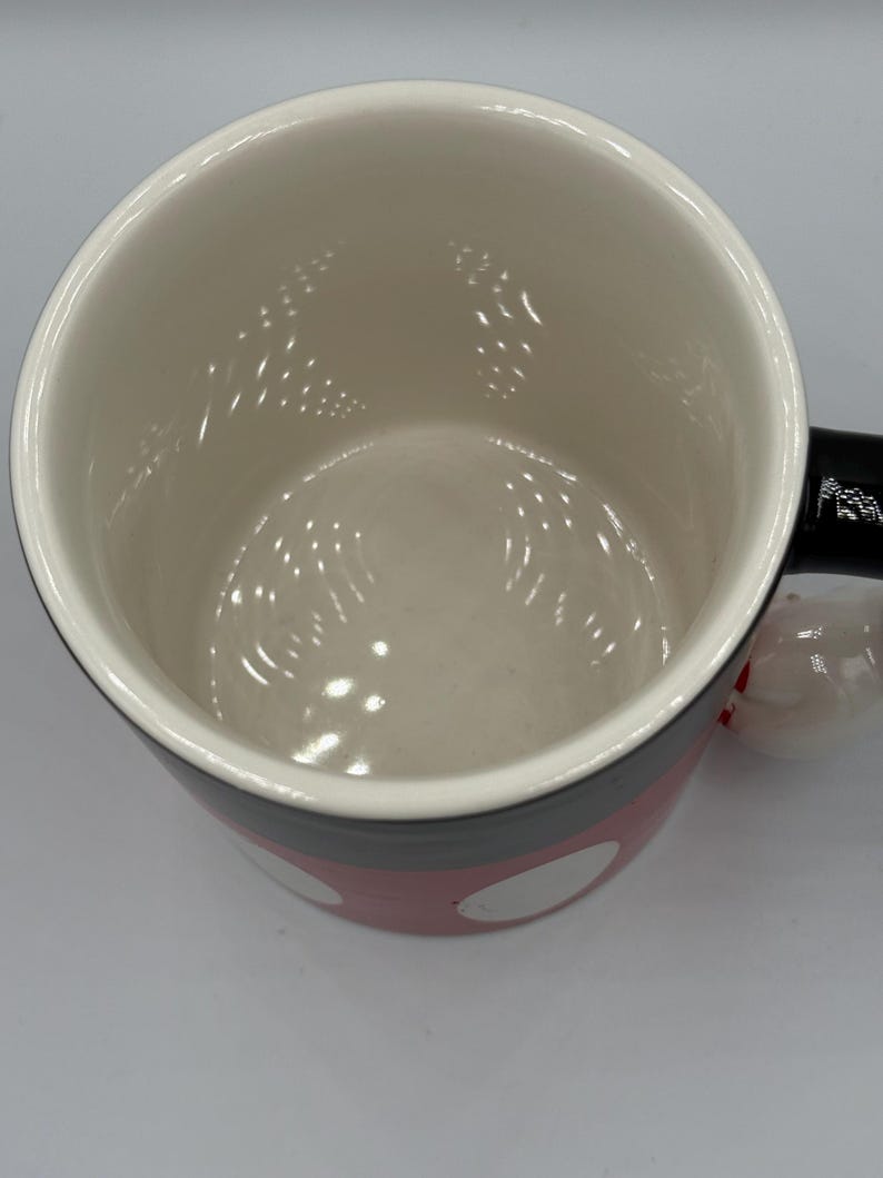 May include: A ceramic mug with a white interior and a red exterior decorated with white polka dots. The mug has a black handle and a black rim. The mug is designed with a cartoon theme.