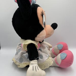 May include: A plush Minnie Mouse doll, featuring black fur, a pink bow with white polka dots, and a light pink face. She wears a polka dot dress with a yellow trim and pink shoes with blue accents. The doll is sitting.