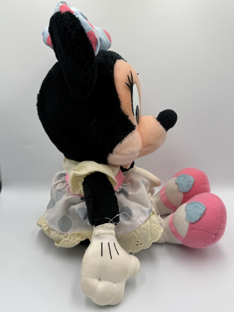 May include: A plush Minnie Mouse doll, featuring black fur, a pink bow with white polka dots, and a light pink face. She wears a polka dot dress with a yellow trim and pink shoes with blue accents. The doll is sitting.