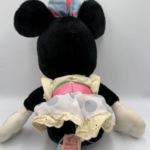 May include: A black plush Minnie Mouse doll with large black ears, wearing a light blue bow with pink polka dots, a white dress with gray polka dots, and a pink belt. The doll has white gloves and a tag.