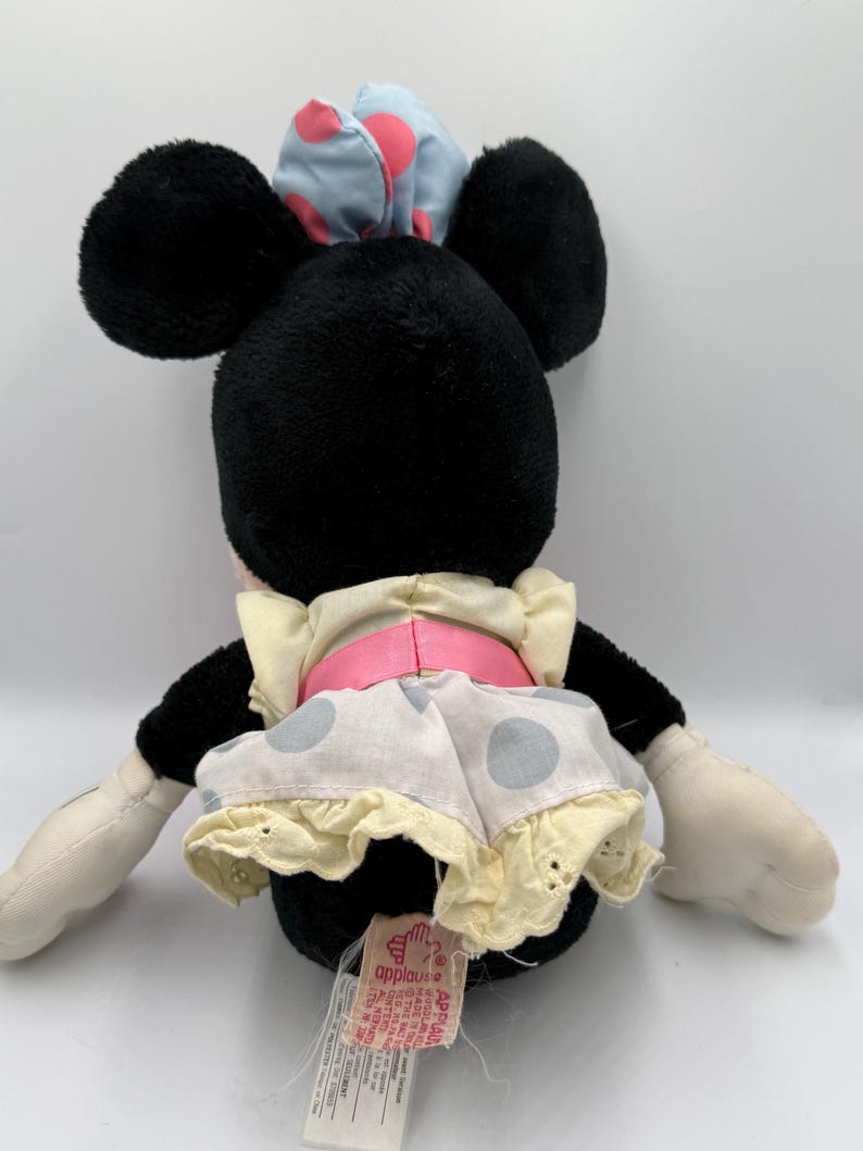 May include: A black plush Minnie Mouse doll with large black ears, wearing a light blue bow with pink polka dots, a white dress with gray polka dots, and a pink belt. The doll has white gloves and a tag.
