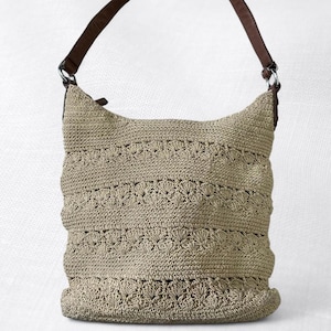 May include: A beige crochet shoulder bag with a brown leather strap. The bag features a detailed, textured pattern and a curved top. The strap is attached to the bag with silver metal rings. The bag is a neutral color and appears to be handmade.