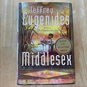 May include: A hardcover copy of the novel "Middlesex" by Jeffrey Eugenides. The cover features an illustration of a figure in a flower, with the title in large white letters. A gold sticker indicates the Pulitzer Prize.