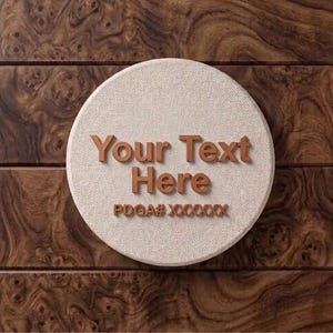 Puede incluir: Una placa circular de tela texturizada en color blanco roto con letras marrones en relieve tridimensional que dicen "Your Text Here" seguidas del texto más pequeño "PDGA# XXXXXX" centrado en la superficie.