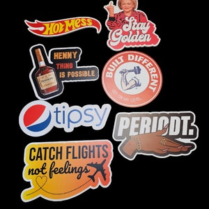 May include: A collection of stickers with various designs and text. One sticker says "Hot Mess" in red and yellow. Another features Betty White with the text "Stay Golden." Other stickers include "Henny Thing Is Possible" and "Catch Flights Not Feelings."