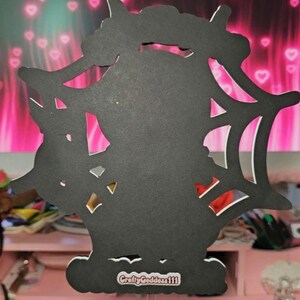 May include: A black, laser-cut decoration featuring a silhouette of a bat within a spiderweb design. The base has a white border and the text "CraftyGoddess111". The background is pink with heart shapes.