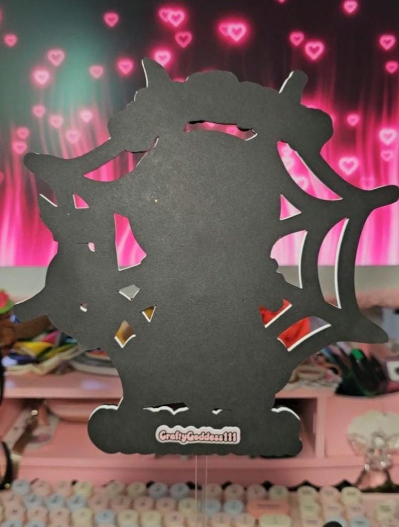 May include: A black, laser-cut decoration featuring a silhouette of a bat within a spiderweb design. The base has a white border and the text "CraftyGoddess111". The background is pink with heart shapes.