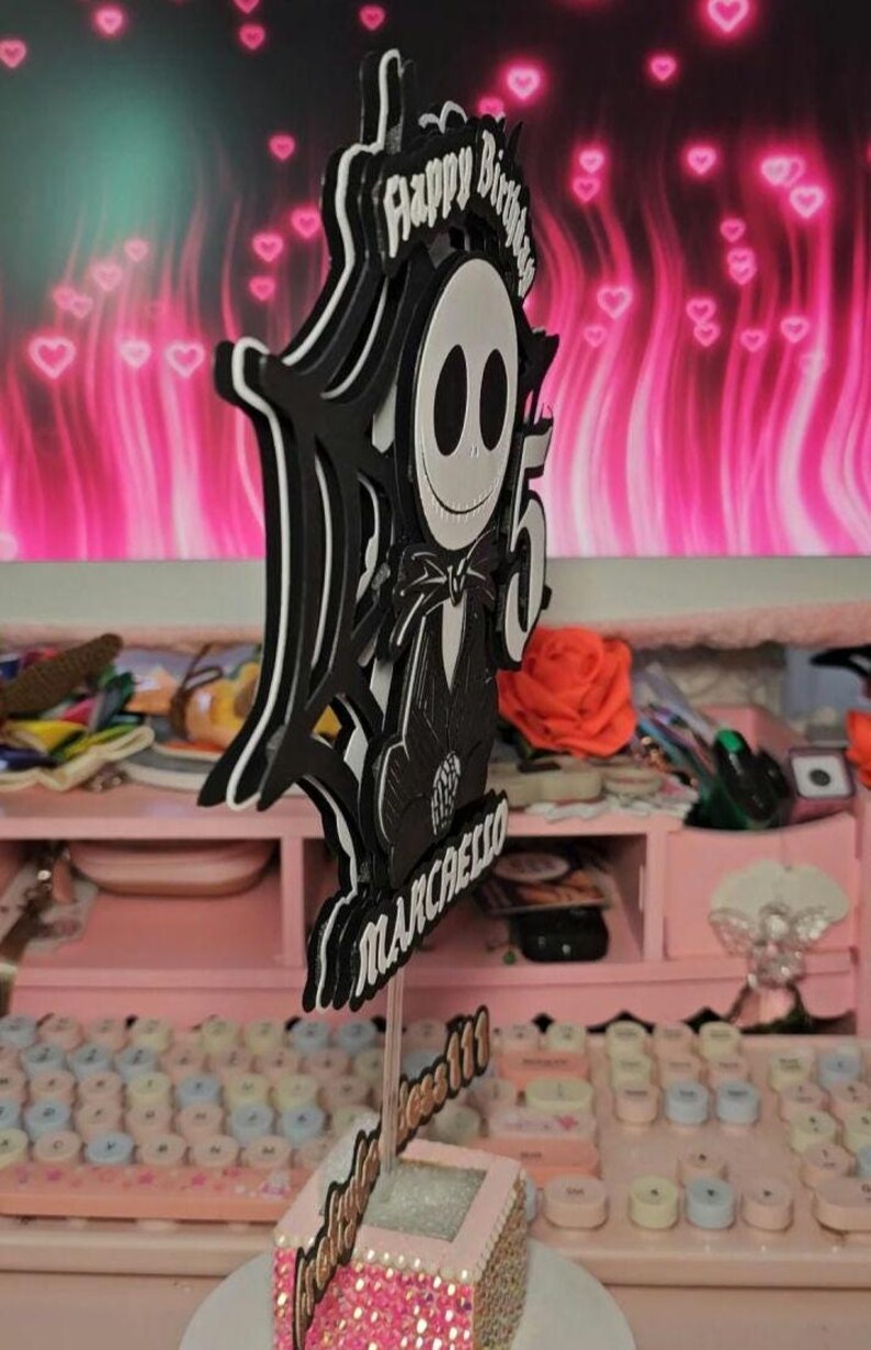 May include: A black and white layered cake topper featuring a cartoon character with the text "Happy Birthday" and the number "5". The name "Marchello" is also included. The base of the topper is pink and glittery.