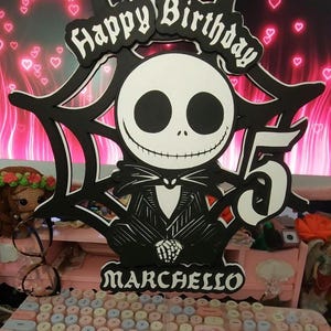 May include: A black and white cake topper featuring Jack Skellington from The Nightmare Before Christmas, with the words "Happy Birthday" and the number "5". The name "Marchello" is also displayed. The topper is set against a pink background.