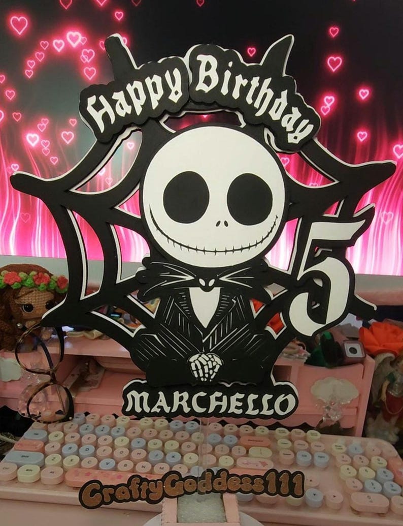 May include: A black and white cake topper featuring Jack Skellington from The Nightmare Before Christmas, with the words "Happy Birthday" and the number "5". The name "Marchello" is also displayed. The topper is set against a pink background.