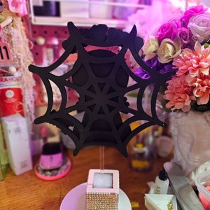May include: A black spiderweb decoration on a wooden stand. The stand has a white base with gold-colored rhinestones and a small pink box with gold rhinestones. The background is blurred with flowers and other decorations.