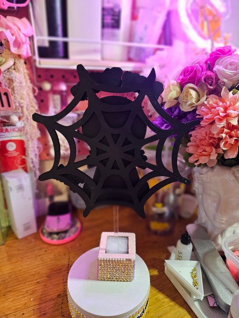 May include: A black spiderweb decoration on a wooden stand. The stand has a white base with gold-colored rhinestones and a small pink box with gold rhinestones. The background is blurred with flowers and other decorations.