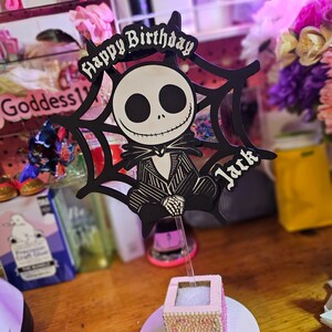 May include: A black and white Jack Skellington cake topper with the words "Happy Birthday Jack" in a gothic font. The topper is set in a small, pink, bedazzled box on a white base, perfect for a themed birthday celebration.