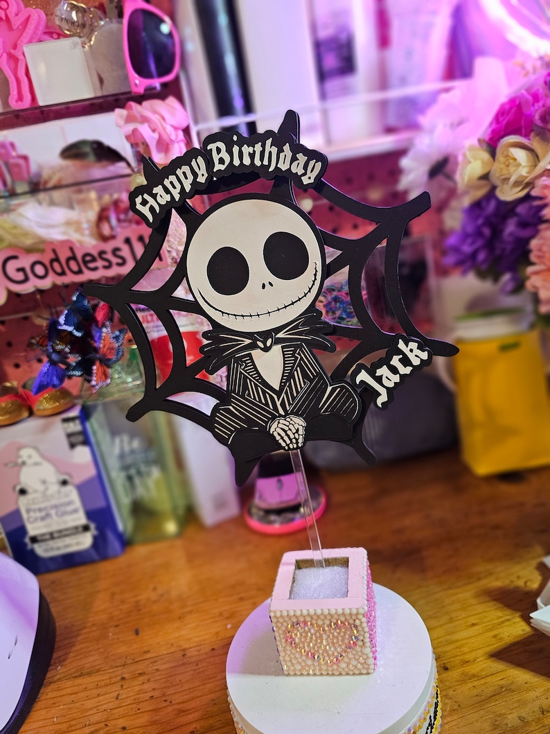 May include: A black and white Jack Skellington cake topper with the words "Happy Birthday Jack" in a gothic font. The topper is set in a small, pink, bedazzled box on a white base, perfect for a themed birthday celebration.